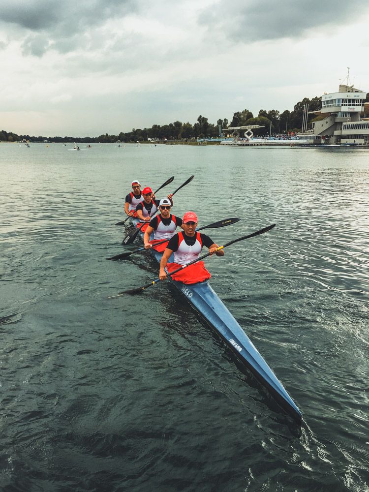 K4 con quattro atleti in canottiera rossa e bianca del CNO e cappellino rosso, sul campo di regata di un lago con tribuna e torre di arrivo