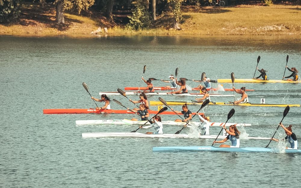Partenza di una gara di K1 femminile, otto kayak affiancati sull'acqua verde con gli alberi sullo sfondo