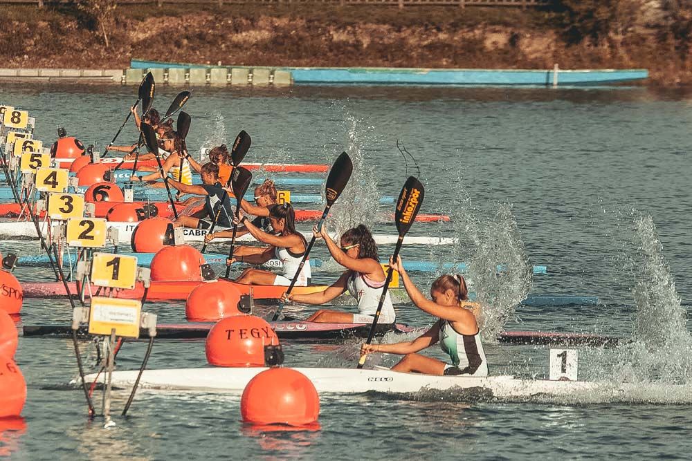 Partenza di K1 femminile dal pontile con i cartellini di corsia da 1 a 8, boe rosse e spruzzi nella luce calda
