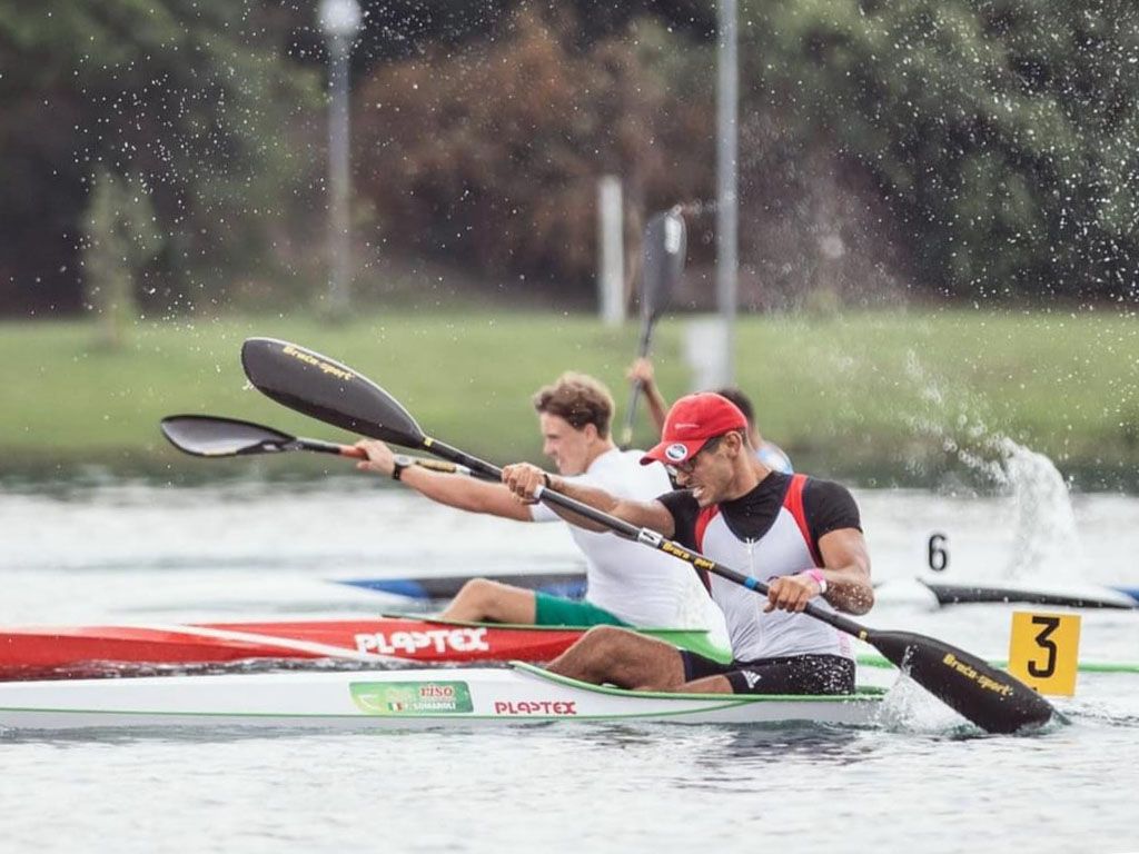 Atleta in K1 con cappellino rosso e canottiera bianca e rossa in piena pagaiata, spruzzi, cartellino di corsia 3
