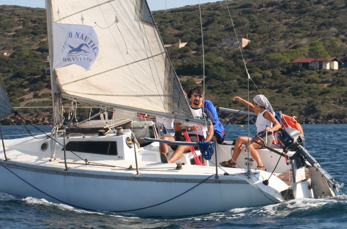 Cabinato a vela con la bandiera del Circolo Nautico Oristano, un uomo al timone e una ragazza a poppa, costa collinare sullo sfondo