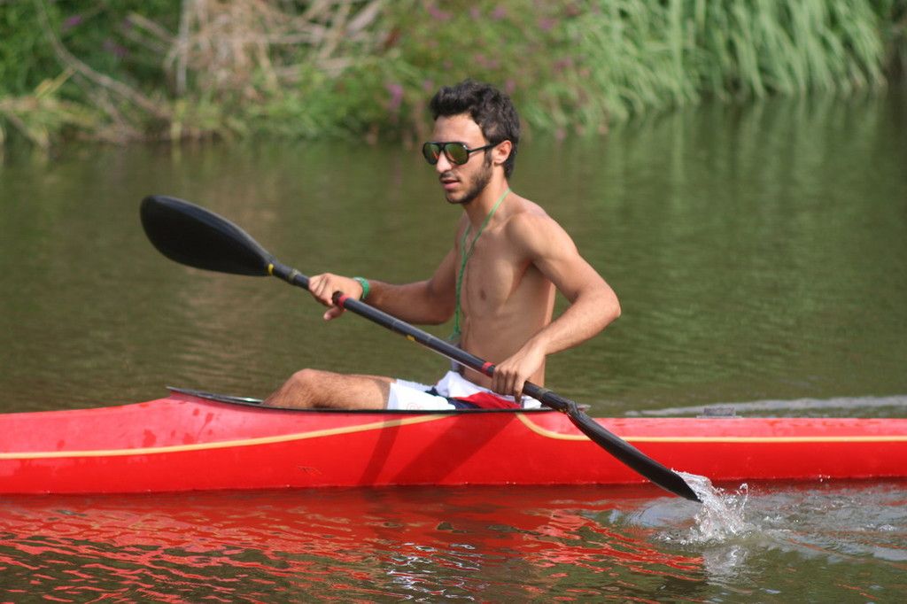 Un ragazzo con gli occhiali da sole pagaia in un kayak rosso, il verde della riva sfocato dietro
