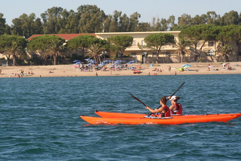 Due ragazze in un surfski doppio arancione con i giubbotti rossi; dietro la spiaggia di Torregrande con gli ombrelloni e i pini