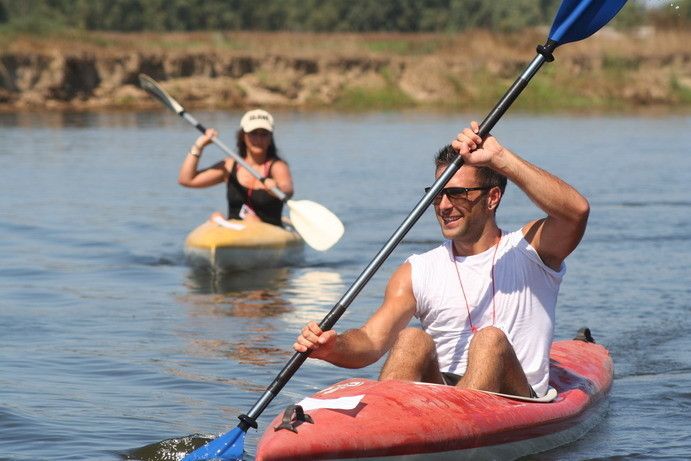 Un uomo in kayak rosso con gli occhiali da sole in piena pagaiata, dietro una ragazza in kayak giallo, argine sabbioso